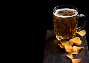 Glass of lager beer with potato crisps snack on vintage wooden board on black background. Beer and snack. Space for text