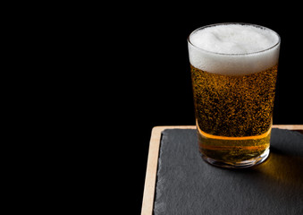 Glass of lager beer with foam and bubbles on stone board on black background. Space for text