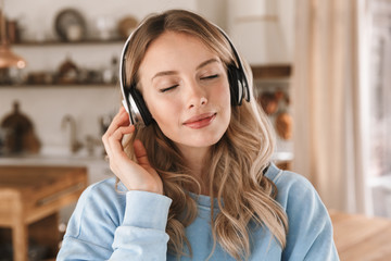 Portrait of content blond girl 20s wearing headphones listening to music at home