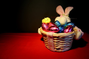 Easter basket with foil eggs on a red cloth. Basket is to the right of the image to allow for text to be placed on the left. There is a small chick resting on an egg in the basket.