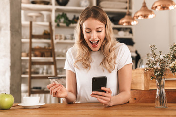 Portrait of gorgeous blond woman using smartphone and plastic credit card while sitting in cozy cafe indoor - Powered by Adobe