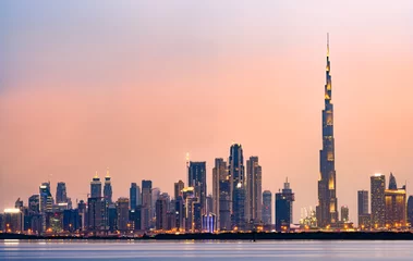 Photo sur Plexiglas Burj Khalifa Vue imprenable sur les toits illuminés de Dubaï au coucher du soleil avec le magnifique Burj Khalifa et de nombreux autres bâtiments et gratte-ciel se reflétant sur une eau douce et soyeuse qui coule au premier plan.