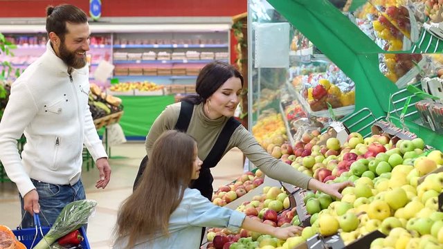 Young parents with a little daughter choose fresh apples in a large supermarket. Happy family buying groceries at the vegetable store. Slow motion.