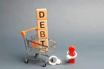 Wooden blocks with the word Debt in a supermarket trolley and a human figurine bound by handcuffs. Mandatory payment of debt. Alimony payment. Pay of a mortgage loan. Debt load / obligation. IOU