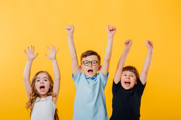 three children, a little boy and two little girls jumping up, their raised hands are happy, isolated yellow background, copy space