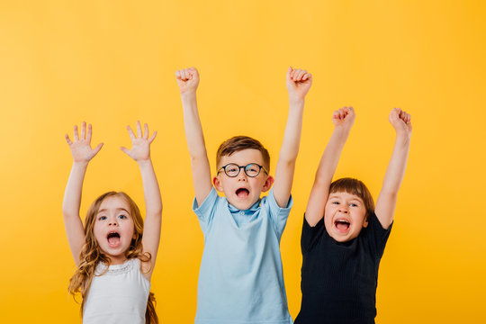 three children, a little boy and two little girls jumping up, their raised hands are happy, isolated yellow background, copy space