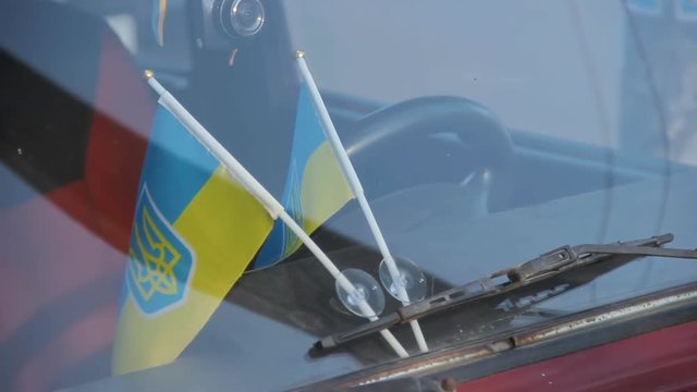 Ukrainian national flags attached to auto windshield glass under car wipers. Old fashioned red colored soviet automobile Zhiguli Vaz Lada in sunny spring day. Ukraine, circa spring 2019