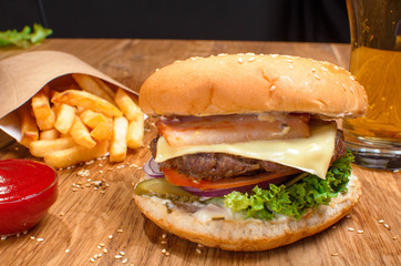Big tasty cheeseburger on the wooden table with french fries. Delicious lunch with cheese, tomatoos and salad. Fast food with beer.