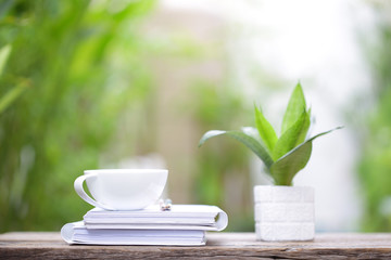 White coffee cup and notebooks with small plant in white pot on brown wooden table at outdoor