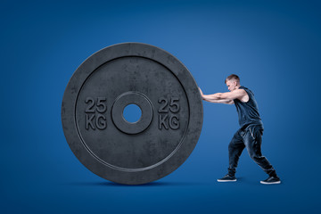 Side view of young athletic little man in sleeveless hoody pushing huge black 25kg weight plate on...