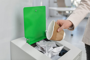 Woman putting used paper cup into trash bin in modern office, closeup. Waste recycling