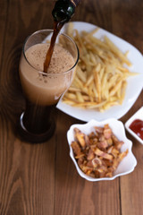 Vertical. The glass is filled with dark beer from the bottle, french fries and snacks in the background. Men's set to watch a football match or a party. Top-side view