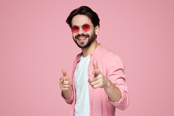Handsome bearded man in stylish shirt and sunglasses showing pointing gesture on pink background