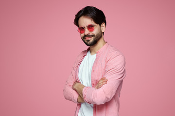Bearded young man keeps hands crossed, wearing pink shirt and glasses, smiling while standing over...