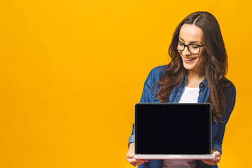 Surprised happy brunette woman in casual showing blank laptop computer screen and pointing on it while looking at the camera with open mouth over yellow background.
