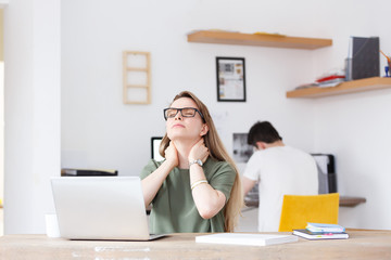 Pain in the neck. Beautiful young woman rubbed tired at the office sitting at table on background of blurred man employee