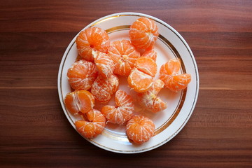 Rounded white plate with lot of orange peeled mandarin (tangerine) citrus. Top view of plate with fruits on the wooden table on kitchen.