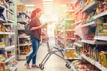 A young beautiful woman rolls a grocery cart and chooses products in the supermarket. Light