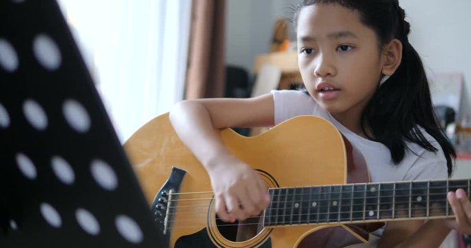 Asian little girl learning to play basic guitar by using acoustic guitar for beginner music instrumental self studying at home