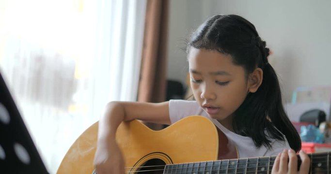 Asian little girl learning to play basic guitar by using acoustic guitar for beginner music instrumental self studying at home