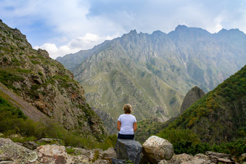 A young woman in the valley among the peaks of Stepantsminda (Kazbegi) sits on the rocks and gazing into the distance. Natural landscape of Georgia.