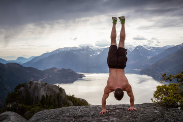 Fit and Muscular Young Man is doing a handstand on top of the Mountain during a cloudy day. Taken on Chief Mountain in Squamish, North of Vancouver, BC, Canada.