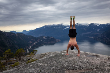 Fit and Muscular Young Man is doing a handstand on top of the Mountain during a cloudy day. Taken on Chief Mountain in Squamish, North of Vancouver, BC, Canada.