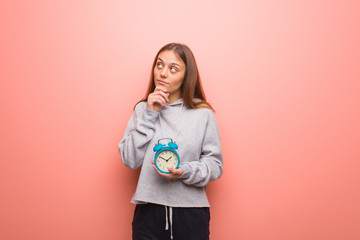 Young pretty caucasian woman doubting and confused. She is holding an alarm clock.