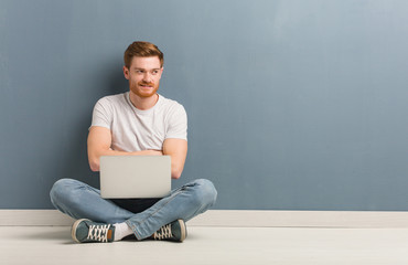 Young redhead student man sitting on the floor smiling confident and crossing arms, looking up. He is holding a laptop.