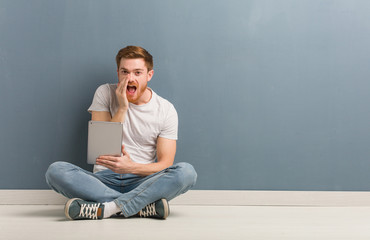 Young redhead student man sitting on the floor shouting something happy to the front. He is holding a tablet.