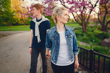 stylish and beautiful woman with short light hair, dressed in a blue jeans jacket walks with her handsome man in a sunny park and standind near flower trees