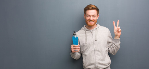 Young redhead fitness man showing number two. He is holding an energy drink.