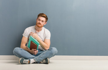 Young redhead student man sitting on the floor smiling confident and crossing arms, looking up. He is holding books.