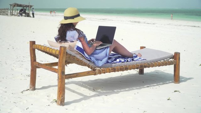 A young woman uses a laptop on a white beach near the ocean. Adult caucasian girl in straw hat and sundress is lying on lounger sunbed on white sand coast line and typing on laptop at bright suny day