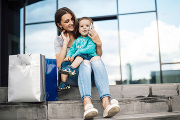 Mother sitting with son and using phone with packages near mall after shopping.