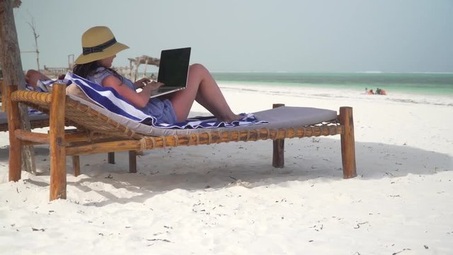 A young woman uses a laptop on a white beach near the ocean. Adult caucasian girl in straw hat and sundress is lying on lounger sunbed on white sand coast line and typing on laptop at bright suny day