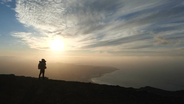 Silhouette of a girl in bicycle helmet with backpack taking pictures of an incredible sunset on the top of a Mountain above the ocean on Canary Islands. Lanzarote.