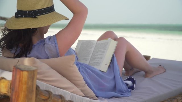 Young woman is reading book on white sand beach by ocean. Adult caucasian girl in straw hat and sundress is lying on lounger sunbed in shadow on white sandy beach and reading book at bright sunny day.