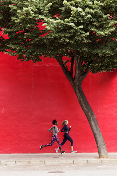 Two sporty young women running together in the city passing red wall