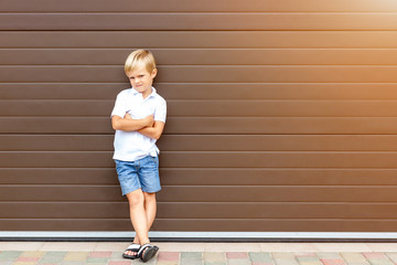Cute grumpy blond child in casual clothing standing against brown garage door. Angry kid boy with...