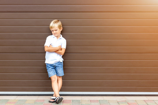 Cute grumpy blond child in casual clothing standing against brown garage door. Angry kid boy with crossed arms near house.Awkward age and parenting concept. Children and parents relations