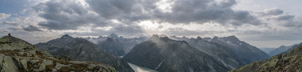 Obraz premium Panoramic view to the Grimsel lake in Swiss Alps with dramatic sun rays breaking through dark clouds with a boy resting on a rock