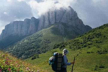 A woman with a backpack in a vest and hat stands on an alpine and looks at the mountain Aceshbok
