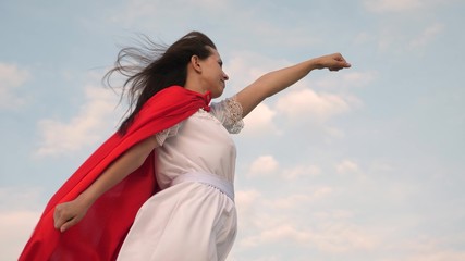Fototapeta na wymiar beautiful superhero girl standing on a field in a red cloak, cloak fluttering in the wind. girl dreams of becoming superhero. Slow motion. young woman plays in a red cloak with expression of dreams.