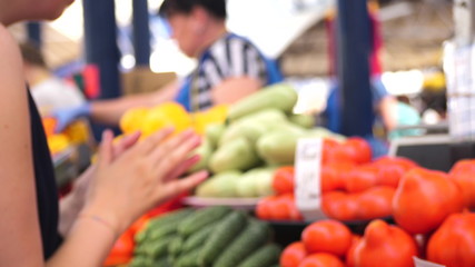 young beautiful woman choosing tomatoes on greenery counter in the city marketplace with an unrecognizable seller on the background, side view close up slow motion video in 4K