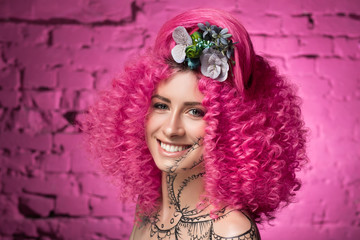 Young attractive caucasian girl model with african style curly bright pink hair, tattooed face and neck and flowers woven into her hair. Photo in the studio on a pink background, smile into the camera