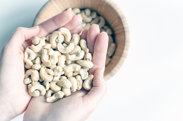 Two women's hands holding a handful of cashew nuts, close-up. On the back blurred plan wooden dish with nuts.