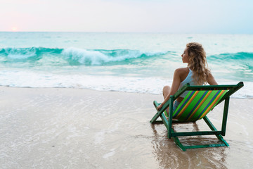 Portrait of beautiful young caucasian woman sitting on sun lounger at beach at sunrise and relaxing. Morning meditation concept