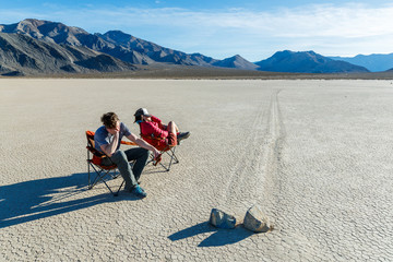 "Exciting day at the races".  Two people sit in chairs asleep while watching the very slow results of two competing boulders at The Racetrack in Death Valley National Park. The area gets its name from the once mysterious tracks which are the result of periodic winter storms over many years that brought moisture to the playa followed by cold night time temperatures which then formed thin sheets of ice and then are pushed by high winds, nudging the boulders a little at a time and creating these remarkable tracks. Disclaimer: The playa is a senstive environment and the site of theft and vandalism. The participants in this photo were careful to make sure the ground was dry and that the chairs left no trace.