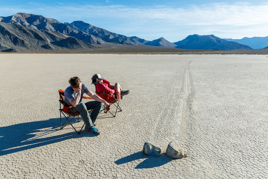 "Exciting day at the races".  Two people sit in chairs asleep while watching the very slow results of two competing boulders at The Racetrack in Death Valley National Park. The area gets its name from the once mysterious tracks which are the result of periodic winter storms over many years that brought moisture to the playa followed by cold night time temperatures which then formed thin sheets of ice and then are pushed by high winds, nudging the boulders a little at a time and creating these remarkable tracks. Disclaimer: The playa is a senstive environment and the site of theft and vandalism. The participants in this photo were careful to make sure the ground was dry and that the chairs left no trace.